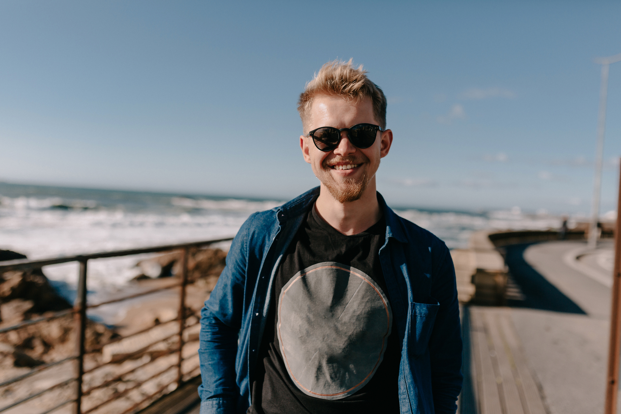 Smiling man with sunglasses and a beard, wearing a dark t-shirt and denim shirt, stands on a boardwalk by the ocean. The background shows waves, rocks, and a clear blue sky.