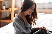 A woman sits on a bed, holding her neck with one hand, and appears to be in pain. She is wearing a gray sweater, with light coming from a window behind her. A wooden shelf is in the background.