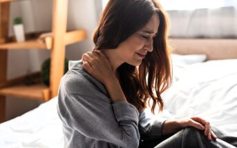 A woman sits on a bed, holding her neck with one hand, and appears to be in pain. She is wearing a gray sweater, with light coming from a window behind her. A wooden shelf is in the background.