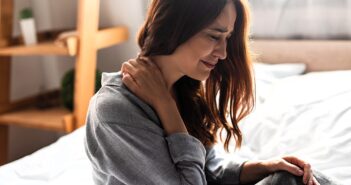 A woman sits on a bed, holding her neck with one hand, and appears to be in pain. She is wearing a gray sweater, with light coming from a window behind her. A wooden shelf is in the background.