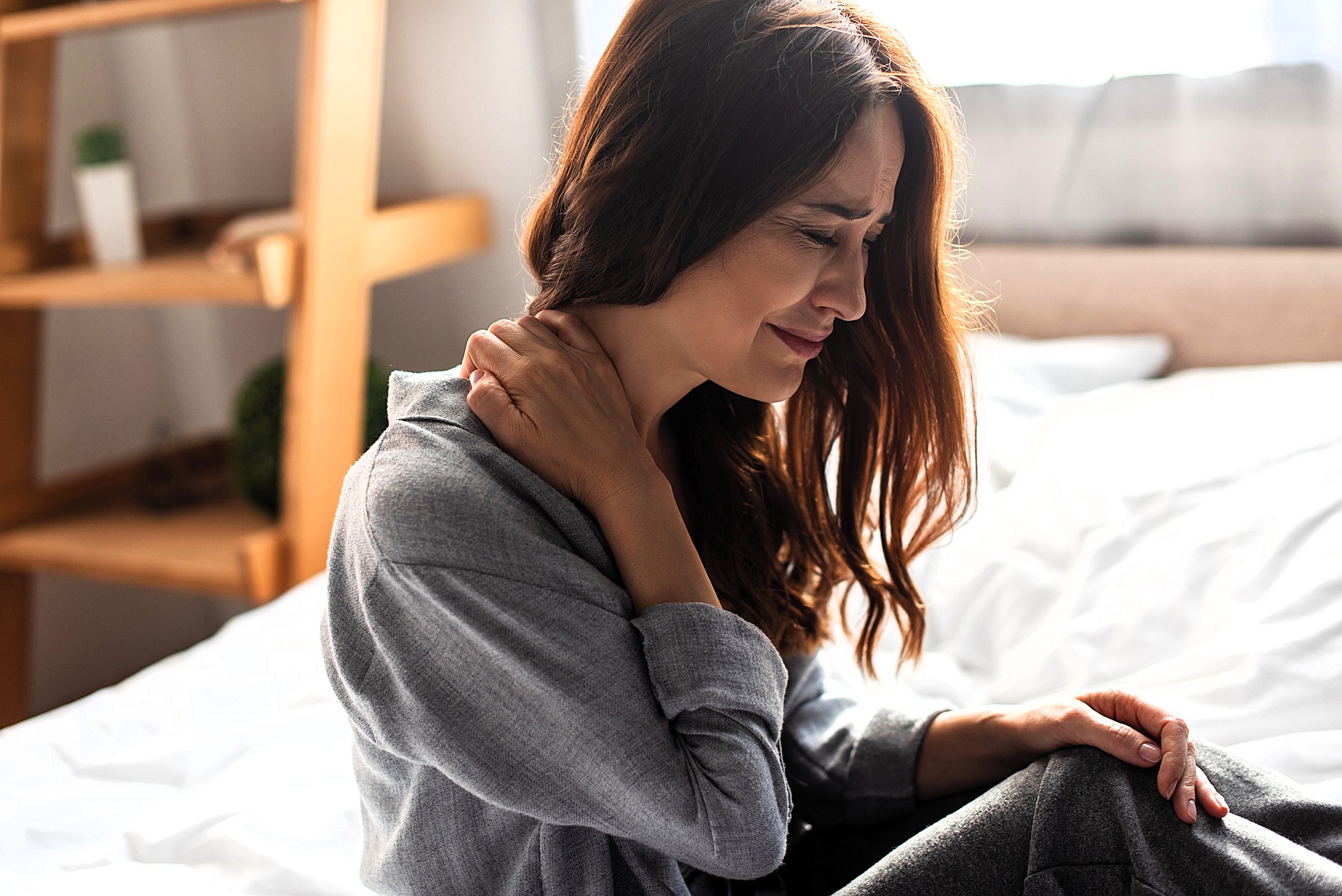 A woman sits on a bed, holding her neck with one hand, and appears to be in pain. She is wearing a gray sweater, with light coming from a window behind her. A wooden shelf is in the background.