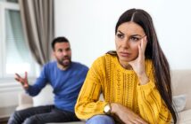 A woman in a yellow sweater sits on a couch looking upset, resting her head on her hand. In the background, a man in a blue sweater is gesturing and appears to be talking to her.