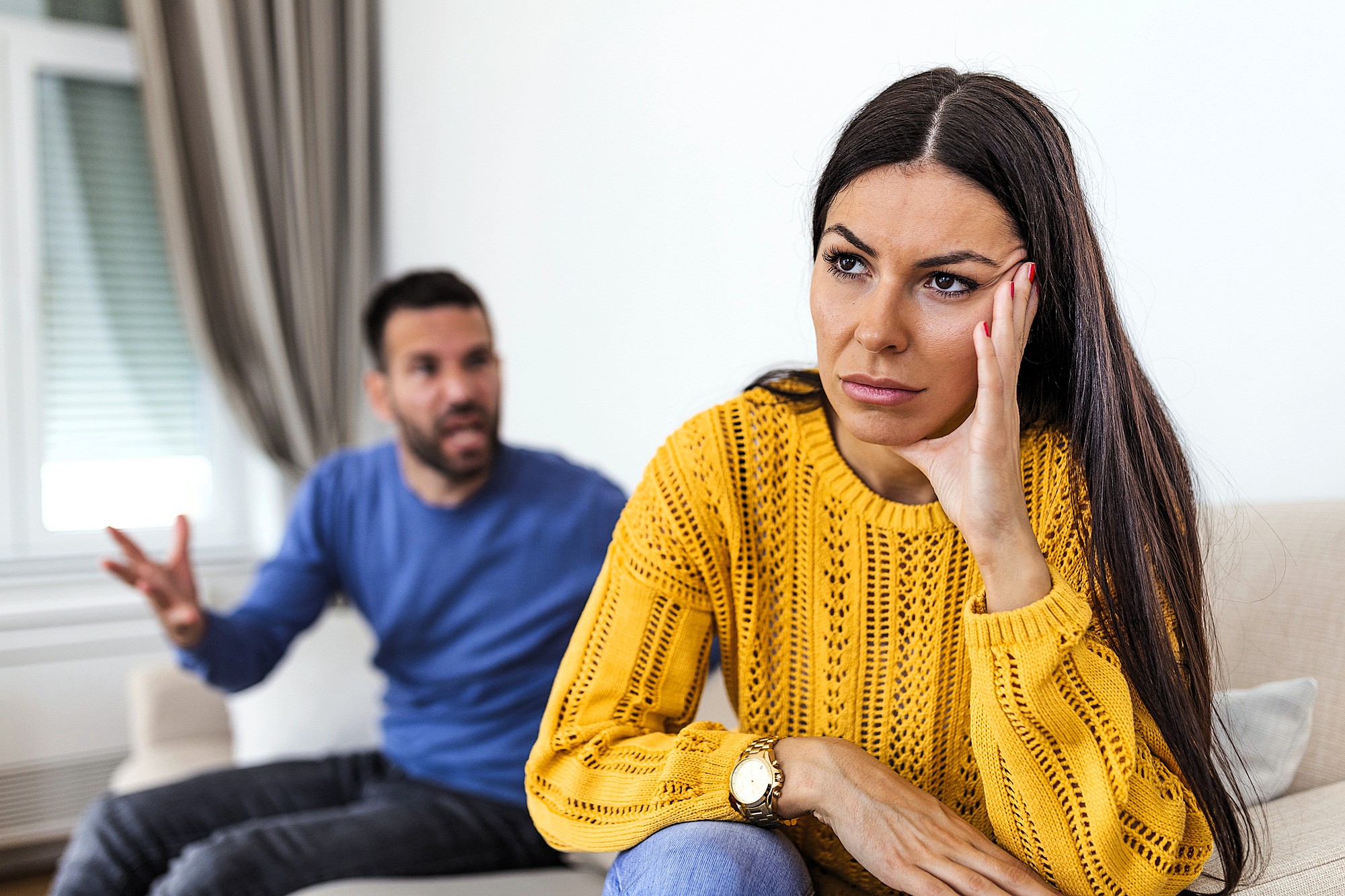 A woman in a yellow sweater sits on a couch looking upset, resting her head on her hand. In the background, a man in a blue sweater is gesturing and appears to be talking to her.