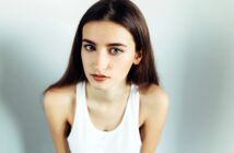 A woman with long brown hair and green eyes is looking directly at the camera. She is wearing a sleeveless white top and is set against a plain background. The lighting highlights her facial features and expression.