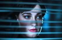 A woman with short dark hair and red lipstick looks through horizontal blinds. Soft blue lighting creates a mysterious atmosphere, highlighting her face and emphasizing her thoughtful expression.