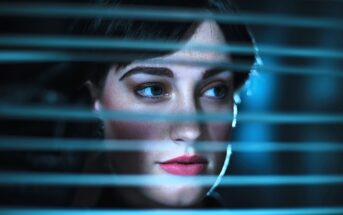 A woman with short dark hair and red lipstick looks through horizontal blinds. Soft blue lighting creates a mysterious atmosphere, highlighting her face and emphasizing her thoughtful expression.