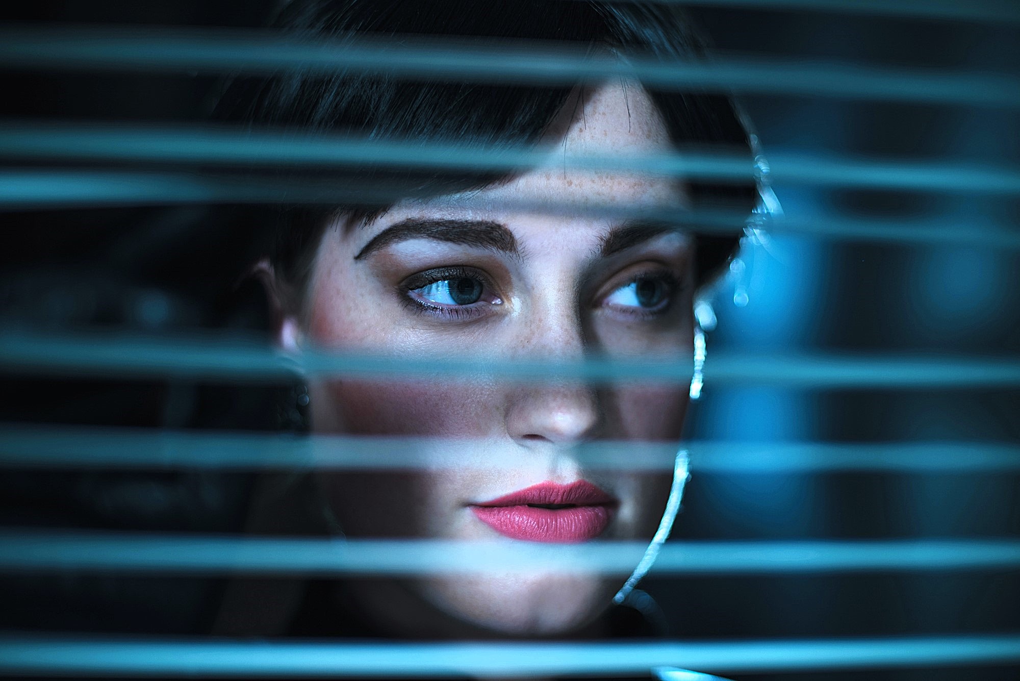 A woman with short dark hair and red lipstick looks through horizontal blinds. Soft blue lighting creates a mysterious atmosphere, highlighting her face and emphasizing her thoughtful expression.