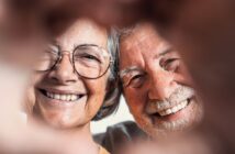 A close-up of a smiling elderly couple. Both are wearing glasses and have gray hair. They are framed by two hands forming a heart shape in the foreground. The couple's expressions are joyful and content.