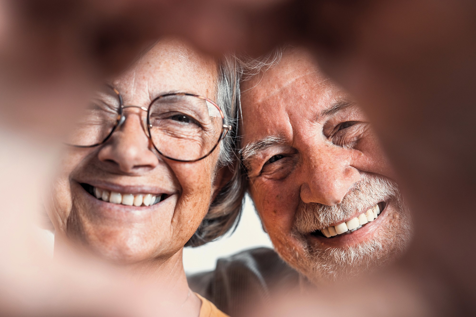 A close-up of a smiling elderly couple. Both are wearing glasses and have gray hair. They are framed by two hands forming a heart shape in the foreground. The couple's expressions are joyful and content.