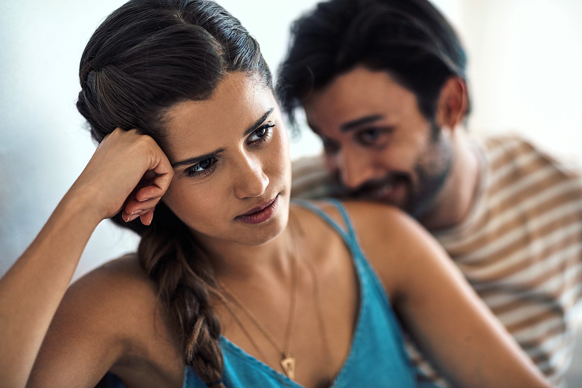 A woman with long braided hair looks thoughtful and slightly upset, resting her head on her hand. A man with a beard and wearing a striped shirt sits closely beside her, appearing to speak animatedly.