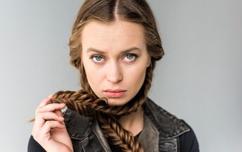 A woman with long braided hair holds part of it in front of her neck. She has green eyes and a serious expression, wearing a black top and a sleeveless denim jacket against a light gray background.