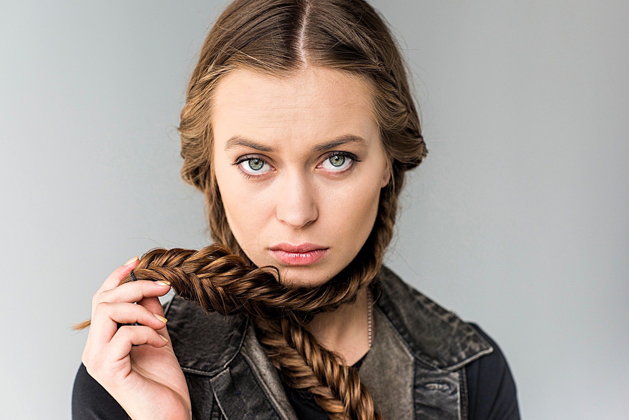 A woman with long braided hair holds part of it in front of her neck. She has green eyes and a serious expression, wearing a black top and a sleeveless denim jacket against a light gray background.